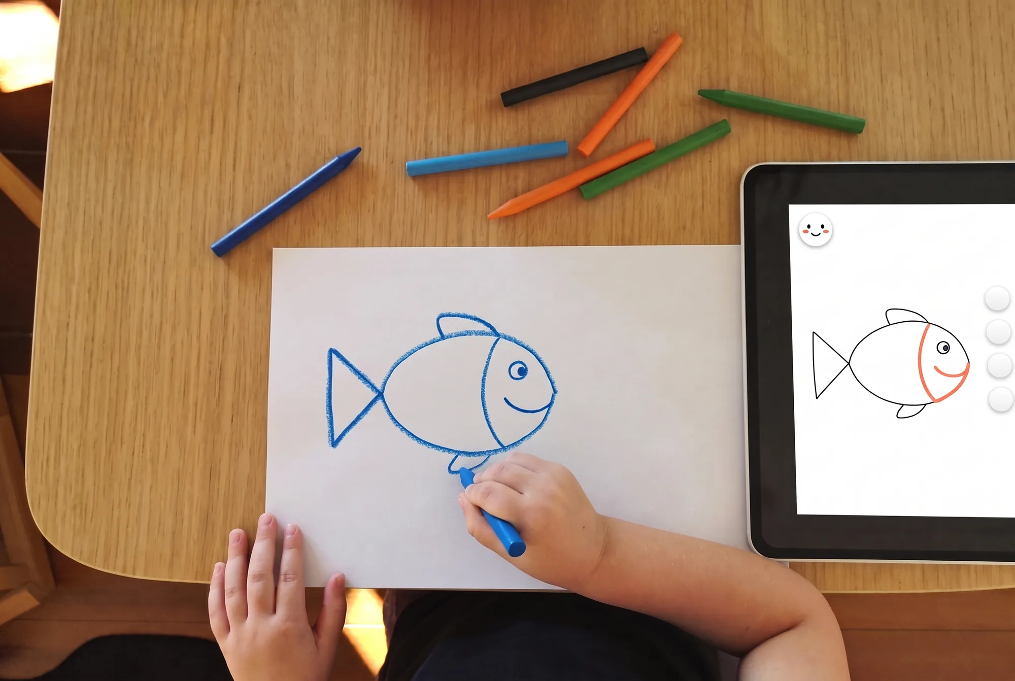 A child’s hands finishing a simple crayon drawing of a fish on paper, with a tablet beside it showing the same fish and the next line highlighted