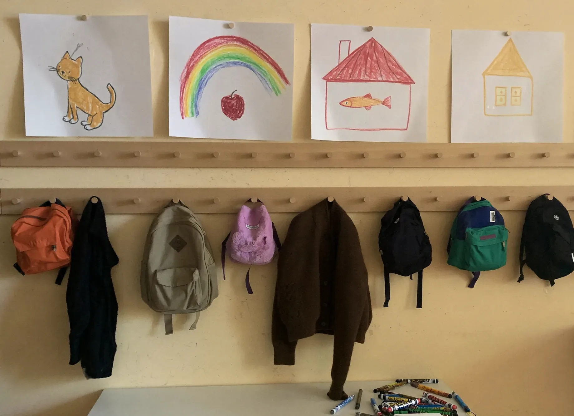 A row of preschool coat pegs on the first morning of term, each with a child’s crayon drawing pinned above it
